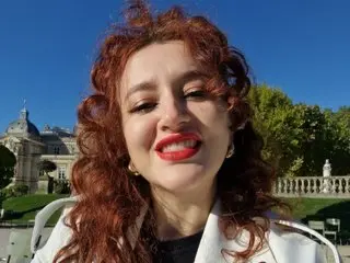 Eva_Jax poses with her long, curly red hair and red lipstick, wearing a white top against a bright outdoor background.