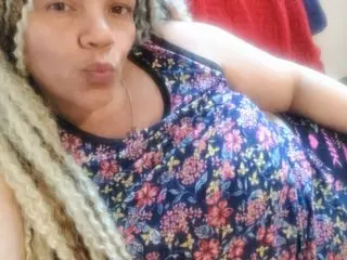 Woman with blonde dreadlocks wearing a blue floral print tank top in a cozy indoor setting.