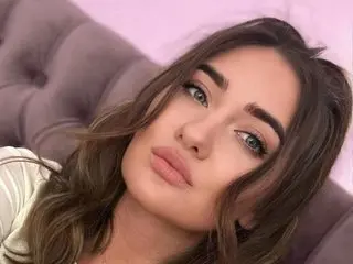 Evalanyss poses for a close-up selfie, showing her face and long brown hair against a soft pink tufted headboard.