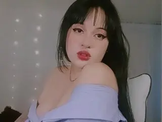 Evamoonn26 poses with long dark hair and red lips, wearing a light purple off-the-shoulder top against a soft, neutral background.