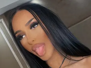 EvePosh poses with her long black hair and tongue out, showing her face and shoulders against a plain background.