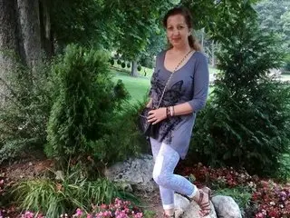 EvelyneFlirtyMilf poses outdoors in a grey long-sleeved top and light blue jeans, standing near green bushes and pink flowers.