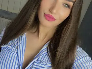Young woman with long brown hair and pink lipstick wearing a blue striped shirt in a bright indoor setting.