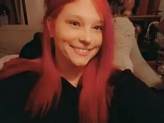 Exoticlexi smiles with her long red hair down, wearing a black top in a dimly lit indoor setting.