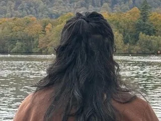 Fabz95 shows her long, dark wavy hair from behind while sitting outdoors near a lake.