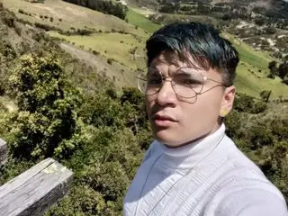 FelipeRey poses outdoors against a lush green hillside, wearing a white turtleneck and looking directly at the camera with short dark hair.