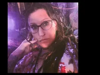 FeralLux33x looks at the camera with a direct gaze, wearing glasses and showing her dark hair and bare shoulders in a close-up shot.