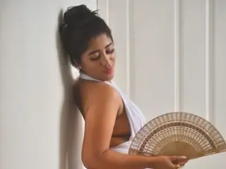 FernandaColonia poses against a white wall, wearing a white top and holding a large decorative fan near her chest.
