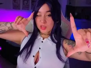 Winking woman with blue hair and tattoos wearing a white sleeveless shirt in a purple neon gaming room.