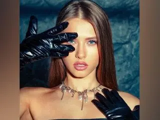 FlexyVelvet poses with her hands framing her face, wearing a silver metal choker against her long brown hair and bare shoulders.