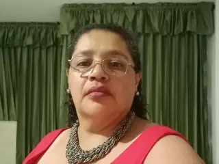 Middle-aged woman with glasses and a silver necklace wearing a red top in front of green curtains.