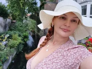 Smiling woman in a pink floral dress and wide-brimmed sun hat relaxing in a lush green garden setting.