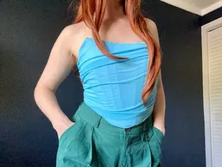 FreyaAvery poses in a light blue tube top and green trousers, showing her long red hair and slim build against a dark background.