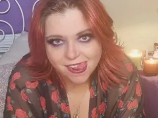 Smiling woman with red hair and smoky eye makeup wearing a floral print robe in a cozy bedroom setting.