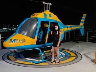GABETAYLOR poses in a black outfit while sitting on a blue and yellow helicopter prop against a dark night background.