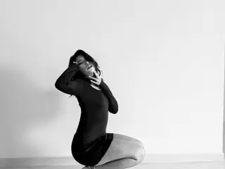 GABRIELAROSI poses in a black long-sleeve bodysuit, tilting her head back with her hands near her face against a plain white background.