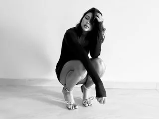 GABRIELAROSI in a black long-sleeve dress crouches on the floor against a white wall.
