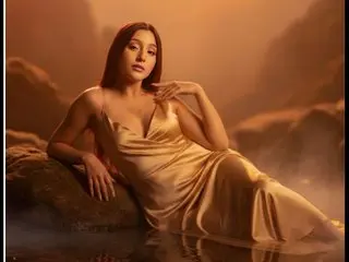 GabbieCarter18 poses in a low-cut gold satin dress, leaning against a dark rock with her hair down in a warm, dim setting.