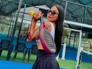 Gabihills poses outdoors wearing a white tank top, holding a tennis racket and a yellow tennis ball against a blue court background.