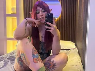 Cosplayer with pink hair and elf ears in green outfit sitting on a bed in a warm lit room.
