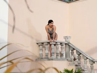 Gema_Hills leans against a white stone banister on a balcony wearing a grey slip dress.
