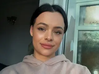 Ghinawilks poses for a close-up selfie, showing her face and shoulders while wearing a light tan hoodie against a window background.