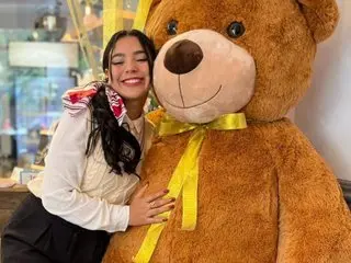 GiaKendric smiles while hugging a large, brown teddy bear, wearing a white long-sleeve shirt and black bottoms with her dark hair down.
