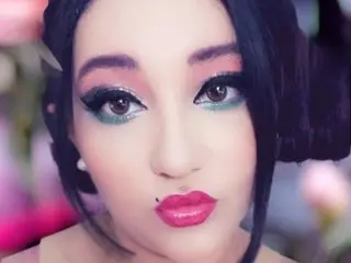 GiaRavenX looks into the camera with heavy eye makeup and red lipstick, showing her face and dark hair against a soft, blurred background.