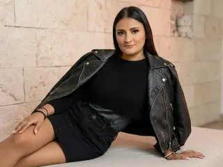 Gia_Smith poses wearing a black leather jacket and black skirt, sitting against a light stone wall with her long dark hair down.