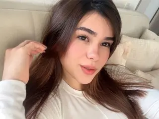 Gianna_Dream poses with her long brown hair pulled back, wearing a white long-sleeve top while looking directly at the camera.