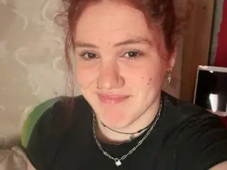 GingerGoddessX is a redhead with a nose ring and silver necklaces wearing a black t-shirt.