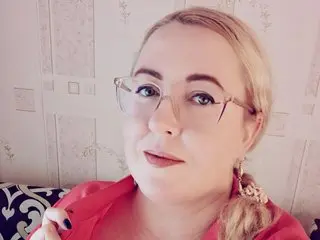 Gingersiya looks at the camera with her blonde hair down, wearing a red top in a room with light patterned wallpaper.