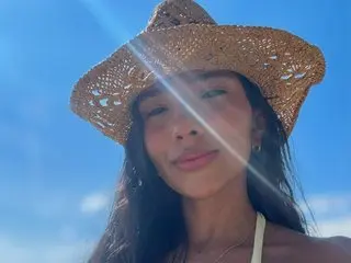 GiselleVera wearing a wide-brimmed straw hat against a clear blue sky, showing her face and long dark hair.