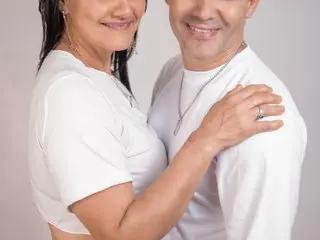 Happy middle-aged couple in matching white outfits sharing a romantic moment against a plain background.