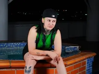 Tattooed woman in a green tank top sitting by a pool at night with a moody expression.