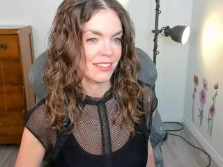 GoddessDelores smiles at the camera with her wavy brown hair while wearing a black mesh top in her room.