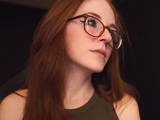 GoddessEmber poses with a close-up view of her face and long red hair, wearing dark-rimmed glasses in a dim setting.