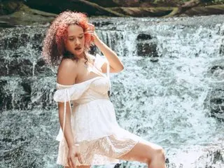 Young woman with curly red hair in a white lace sundress posing by a scenic forest waterfall.