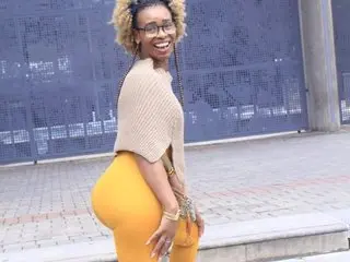 GorillaGripCunt77 smiles while posing in a tan knit crop top and tight yellow leggings, showing her blonde hair and glasses in an outdoor setting.