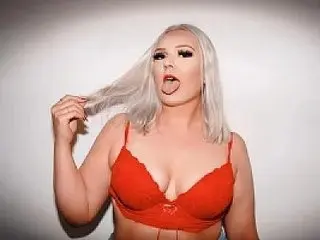GraceLovesBabestation is a blonde woman in a red lace bra posing against a white wall while touching her hair.