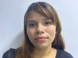 Young woman with long brown hair wearing a black top against a plain white wall background.