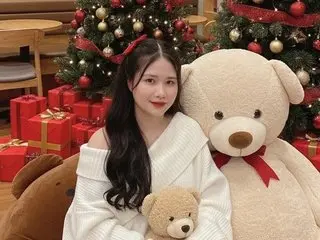 Smiling young woman in a white sweater sitting by a Christmas tree with large teddy bears in a festive setting.