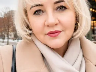 Greatcharisma poses with a soft smile, showing her blonde hair and light eyes while wearing a tan coat and a matching scarf.