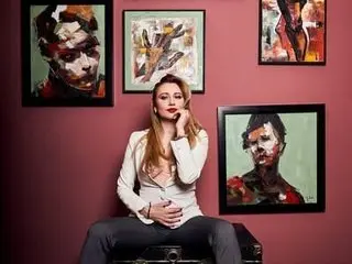 HAILEEYIx poses with her hands near her face, wearing a white long-sleeve top and dark pants against a solid maroon background.