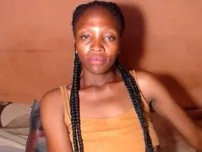 HAIRYSANDY poses looking at the camera with her long black braids hanging down over her shoulders against a plain wall.