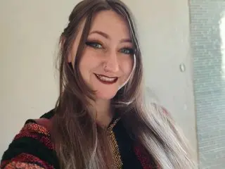 Smiling woman with long hair and dark makeup wearing a colorful patterned bohemian cardigan indoors.