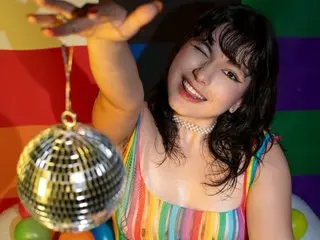 HairyValeri smiles and winks while holding a disco ball in front of a colorful background.