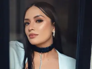 HanaMartinez poses with long dark hair and a black choker, wearing a white top against a dark background.