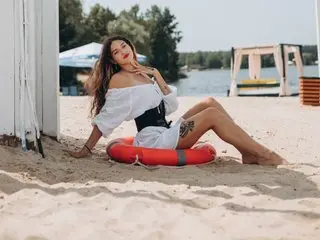 HappyFlowerr poses on a sandy beach wearing a white off-the-shoulder top and black undies, showing her long dark hair and legs.