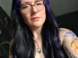 Harley_MoonX is a woman with long dark hair and purple highlights, wearing glasses and a necklace while looking at the camera.